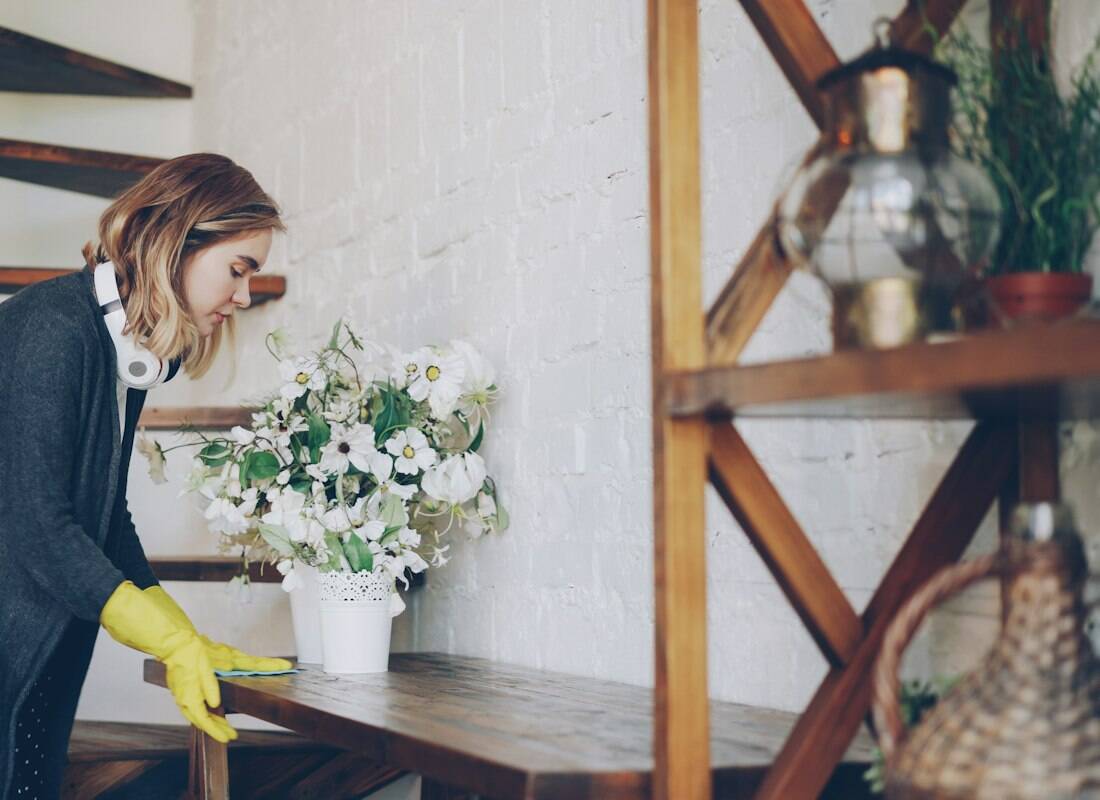 A cleaner in yellow gloves dusting a wooden shelf of plants and glassware.